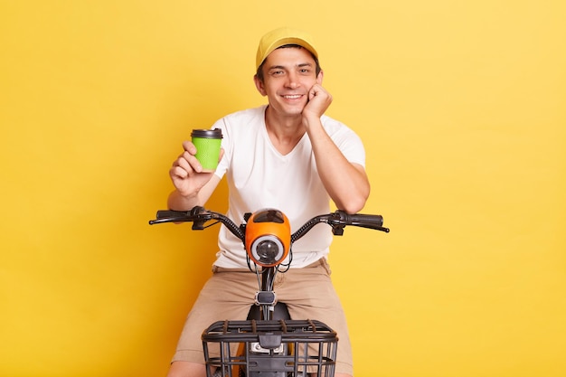 Image d'un homme satisfait portant un t-shirt blanc et une casquette faisant du scooter électrique et s'arrêtant pour boire du café à emporter profitant de son week-end isolé sur fond jaune