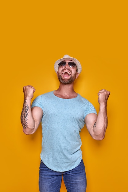 Image d'un homme tatoué barbu excité dans des lunettes de soleil et un chapeau faisant un geste de gagnant isolé jaune ba...