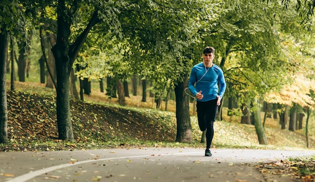 Image horizontale complète du corps d'un jeune jogger faisant du jogging en plein air dans le fond de la forêt Homme de remise en forme faisant de l'exercice dans le parc portant des vêtements de sport bleus et noirs Concept de personnes et de sport