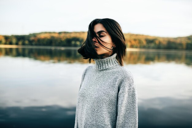 Image horizontale de la magnifique jeune femme brune aux yeux fermés avec les cheveux soufflant portant un pull gris posant sur fond de nature Jolie fille posant contre le lac dans le parc