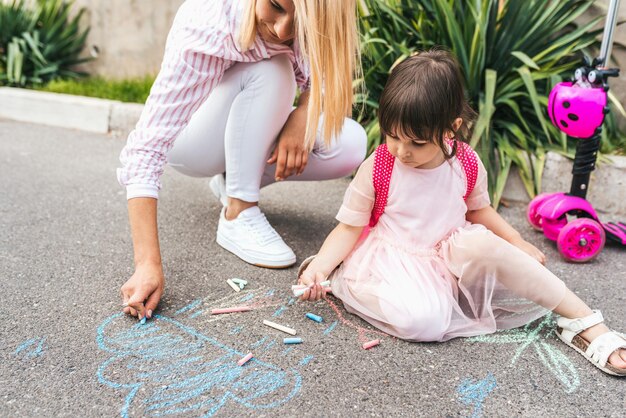 Image horizontale d'une petite fille heureuse et d'une mère dessinant avec des craies sur le trottoir Une femme caucasienne joue avec un enfant d'âge préscolaire avec un sac à dos en plein air Maman et activité enfant Bonne relation