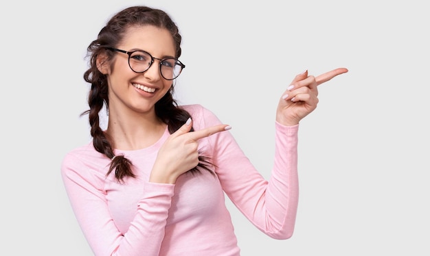 Image intérieure de l'heureuse jeune femme brune à lunettes transparentes indique de côté avec les index à l'espace de copie vierge pour vous texte promotionnel isolé sur mur blanc