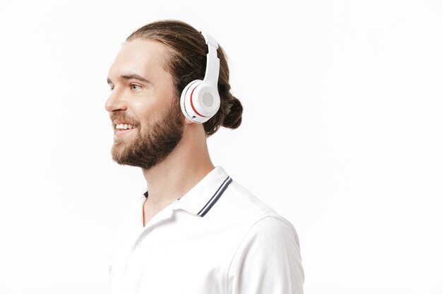 Image d'un jeune bel homme barbu joyeux et joyeux posant isolé sur un mur blanc, écoutant de la musique avec des écouteurs.