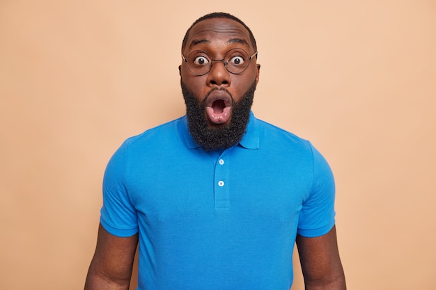 L'image d'un jeune bel homme choqué avec une barbe épaisse regarde des yeux égarés entend de mauvaises nouvelles inattendues porte des lunettes rondes t-shirt bleu isolé sur un mur beige