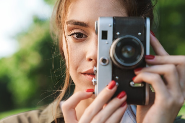 Image d'une jeune brune charmante prenant une photo sur un appareil photo vintage rétro en marchant dans un parc verdoyant par une journée ensoleillée