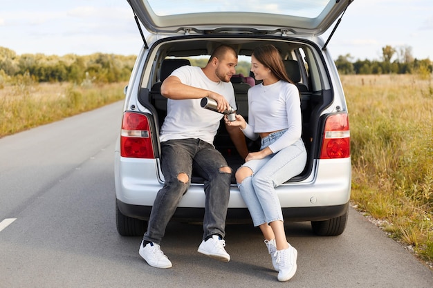 Image d'un jeune couple aimant buvant du thé chaud d'une famille thermos portant des vêtements élégants profitant d'un voyage ensemble s'arrête pour pique-niquer tout en voyageant