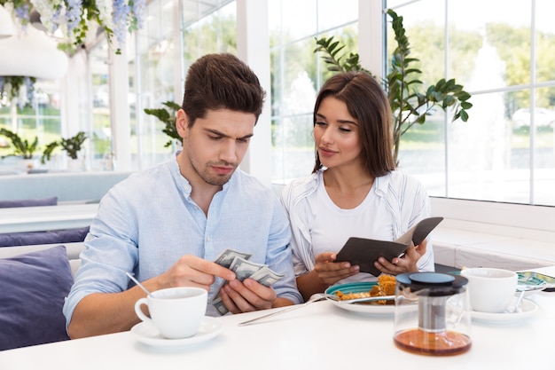 Image d'un jeune couple d'amoureux assis dans un café tenant un chèque et de l'argent.