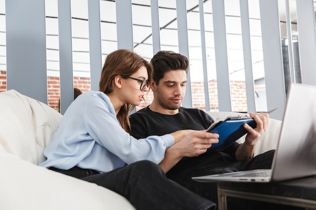 Image d'un jeune couple d'amoureux concentré à la maison à l'intérieur à l'aide d'un ordinateur portable travaillant avec des documents.