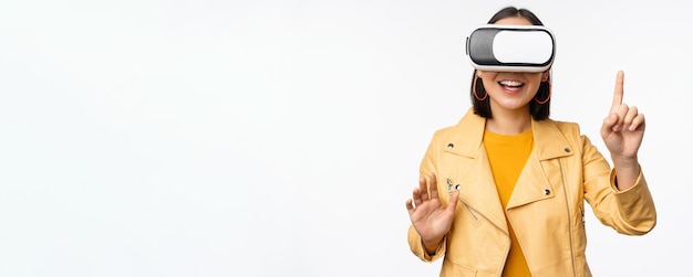 Image d'une jeune femme asiatique portant un casque VR à l'aide de lunettes de réalité virtuelle et souriant pointant vers qch choisissant qch debout sur fond blanc