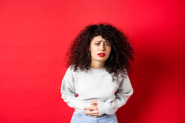 Image d'une jeune femme ayant mal au ventre, se penchant de douleur et se plaignant de crampes menstruelles douloureuses, debout sur fond rouge.