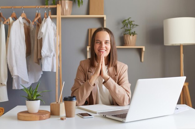 Image d'une jeune femme brune adulte souriante et positive portant une veste beige gardant les mains dans le geste de prière pratiquant le yoga au travail assise sur son lieu de travail devant un ordinateur portable