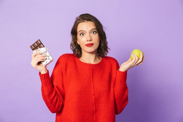 Image d'une jeune femme confuse posant isolé sur mur violet tenant le chocolat et la pomme.