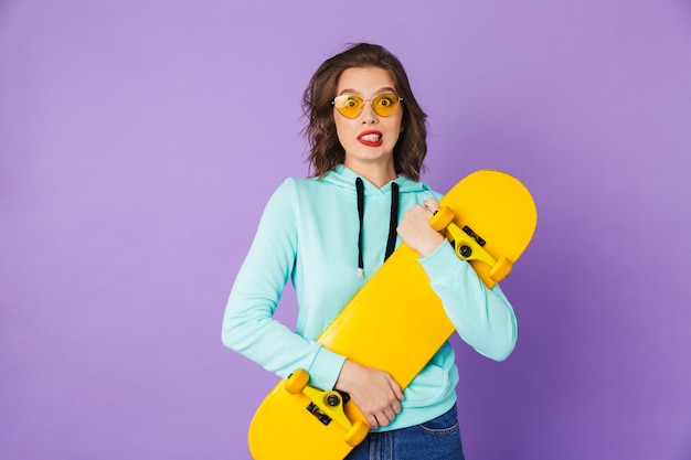 Image d'une jeune femme confuse posant isolée sur mur violet tenant la planche à roulettes.