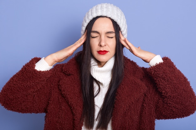 Photo image de jeune femme déçue bouleversée souffrant de maux de tête, mettant les mains sur les tempes, fermant les yeux, ressentant de la douleur, étant seul, debout isolé sur un mur lilas