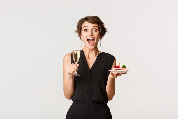 Image de jeune femme étonnée assister à la fête d'anniversaire, tenant un verre de champagne et un gâteau.