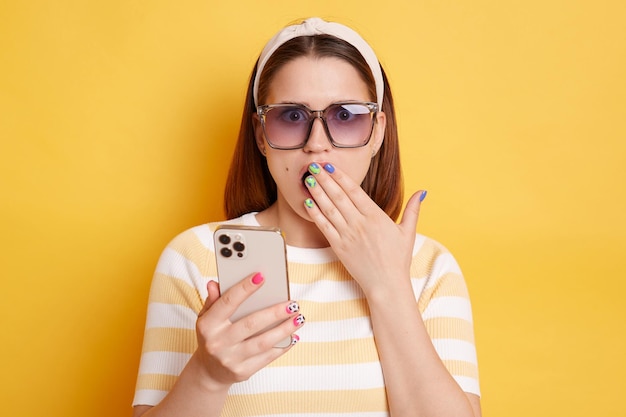 Image d'une jeune femme étonnée choquée portant un t-shirt rayé à l'aide d'un téléphone intelligent lisant des nouvelles choquantes étant surprise de couvrir la bouche posant isolé sur fond jaune