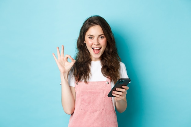 Image d'une jeune femme excitée et heureuse qui dit oui après avoir utilisé l'application pour smartphone, fait des achats en ligne avec un téléphone portable et montré le signe OK, debout sur fond bleu.