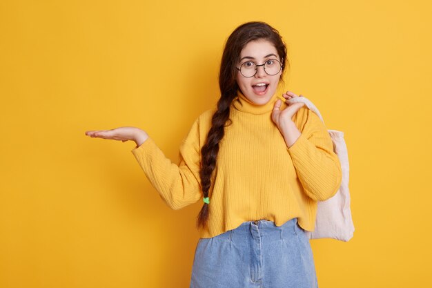 Image d'une jeune femme avec une expression stupéfaite, garde la bouche ouverte, porte une chemise élégante et un jean court, écarte la paume de la main, semble surprise et étonnée.