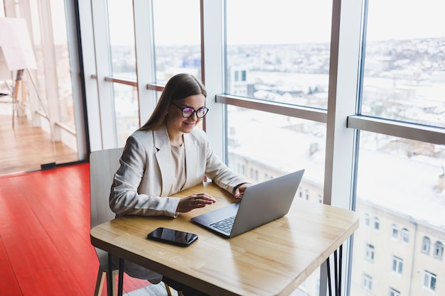 Image d'une jeune femme heureuse dans une veste souriant et travaillant sur un ordinateur portable tout en parlant au téléphone dans un bureau moderne avec de grandes fenêtres