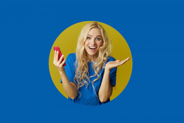 Image De Jeune Femme Heureuse, Debout Sur Un Mur Bleu. Femme Peep Hors D'un Trou Dans Le Mur. Regardant Sur Le Côté Pour Prendre Un Selfie Sur Le Téléphone Mobile.