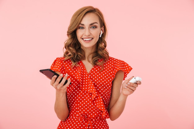 Image d'une jeune femme joyeuse et positive posant isolée sur un mur rose, écoutant de la musique avec des écouteurs bluetooth sans fil à l'aide d'un téléphone portable.