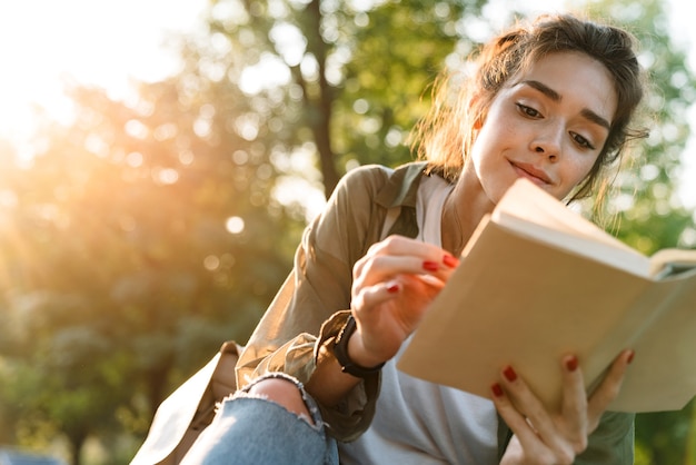Image d'une jeune femme portant des vêtements décontractés souriant et lisant un livre en marchant dans un parc verdoyant