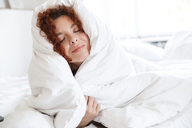 L'image d'une jeune femme rousse ravie en lingerie à l'intérieur d'un hôtel à domicile se trouve dans un lit confortable.