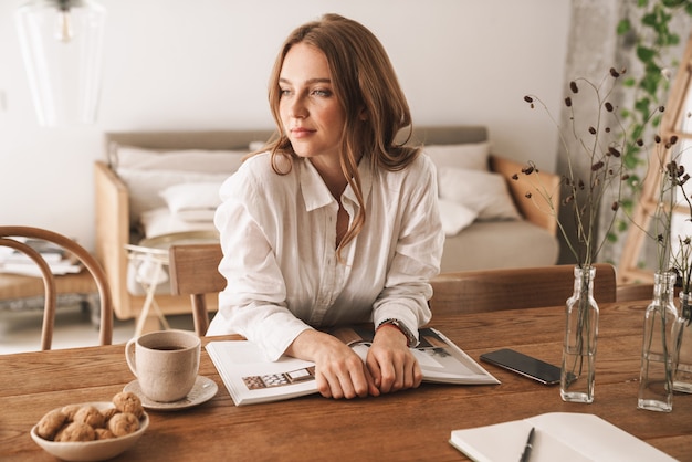 Image d'une jeune femme sérieuse et belle assise à l'intérieur dans un bureau lisant un magazine en regardant de côté.