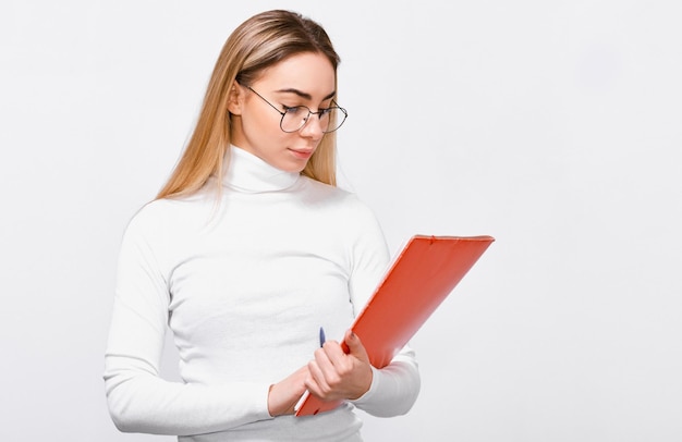Image d'une jeune femme sérieuse portant un chemisier blanc et des lunettes rondes transparentes avec un dossier rouge dans les mains écrivant quelques avis Employée de bureau européenne posant sur fond de studio blanc