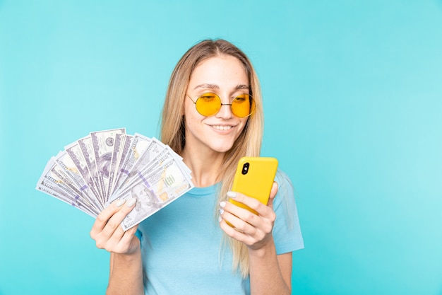 Image de jeune femme souriante tenant le téléphone portable et les billets d'argent isolés sur bleu.