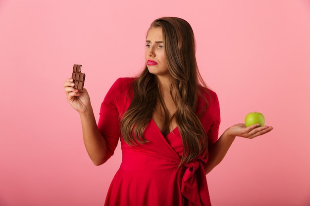 Image d'une jeune femme triste isolée sur un mur rose tenant du chocolat et de la pomme.