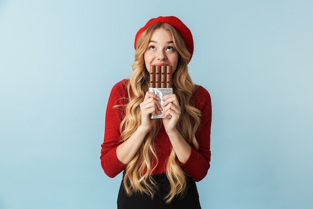 Photo image d'une jeune fille blonde de 20 ans portant un béret rouge mangeant une barre de chocolat isolée