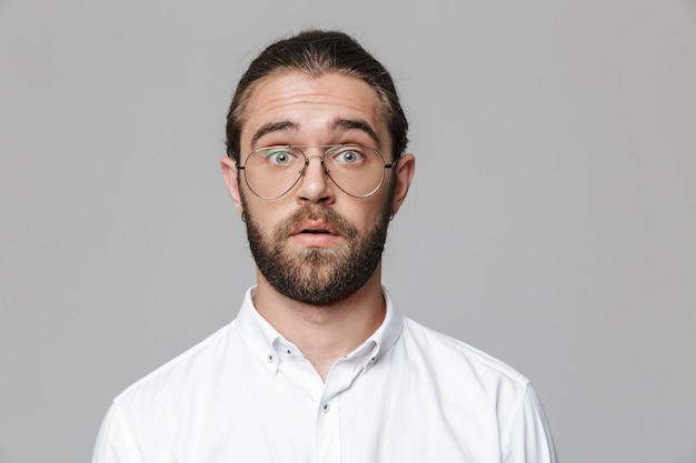 Image d'un jeune homme barbu beau et choqué posant isolé sur un mur gris portant des lunettes.