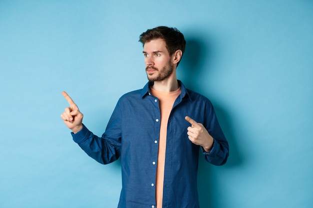 Image de jeune homme caucasien regardant et pointant de côté, montrant la publicité, debout sur fond bleu.