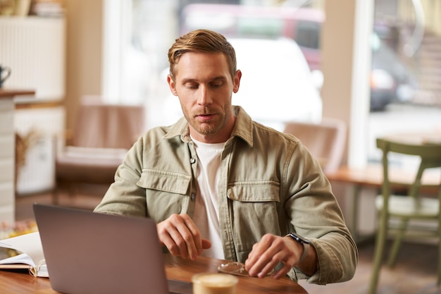 Image d'un jeune homme concentré sur sa tâche assis avec un ordinateur portable dans un café en mettant des lunettes