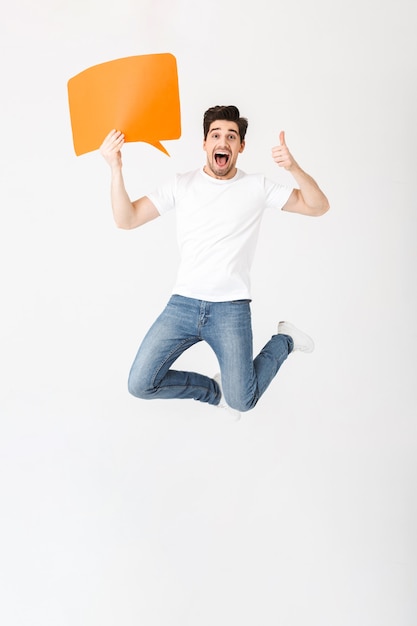 Image d'un jeune homme excité émotionnel posant isolé sur un mur blanc tenant une bulle de dialogue sautant en montrant les pouces vers le haut.