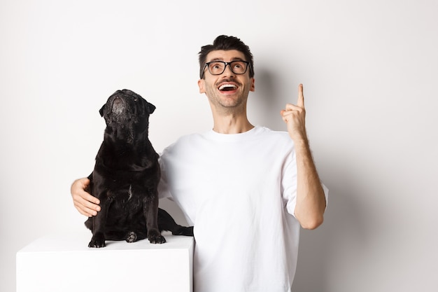 Image d'un jeune homme heureux et d'un chien regardant tous les deux la promo, le propriétaire pointant le doigt vers le haut, le carlin regardant loog, debout sur fond blanc.