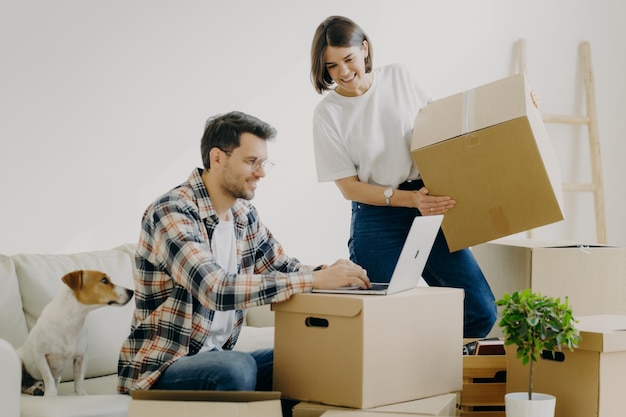 Image d'un jeune homme heureux claviers quelque chose sur un ordinateur portable a une expression heureuse assis sur un canapé confortable avec un animal de compagnie entouré de boîtes en carton faire des achats en ligne acheter de nouveaux meubles