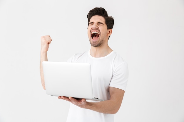 L'image d'un jeune homme heureux et excité posant isolé sur un mur blanc à l'aide d'un ordinateur portable fait un geste gagnant.