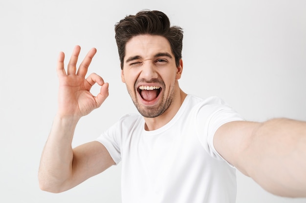 Image d'un jeune homme heureux et excité posant isolé sur un mur blanc fait un selfie par caméra montrant un geste correct.