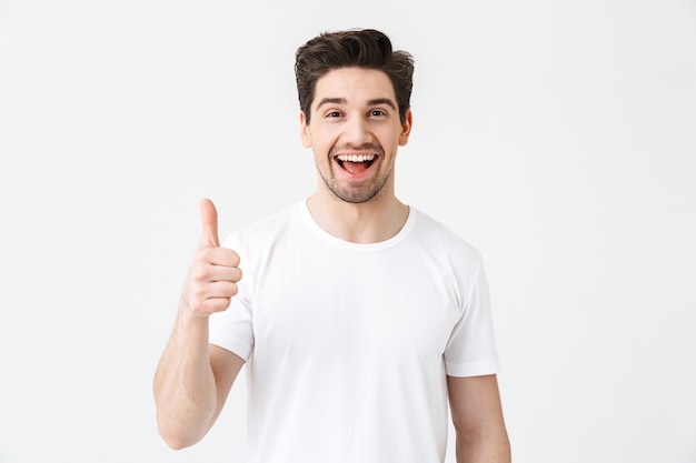 Image d'un jeune homme heureux et excité posant isolé sur un mur blanc montrant le geste du pouce vers le haut.