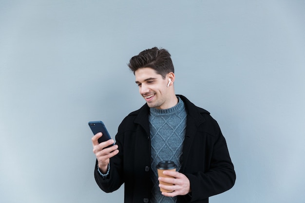 Image d'un jeune homme masculin portant des écouteurs tenant un café à emporter et un téléphone portable tout en se tenant isolé sur un mur gris