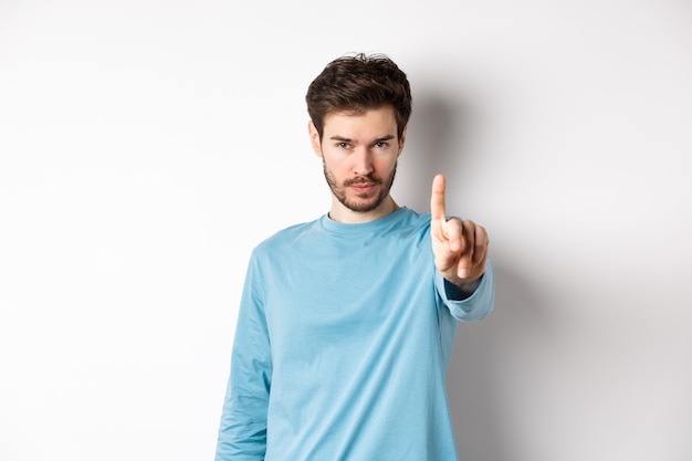Image d'un jeune homme sérieux avec barbe, secouant le doigt en désapprobation, interdire ou interdire quelque chose, debout sur fond blanc et dire non