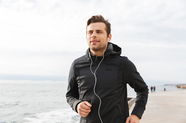 Image d'un jeune sportif de 30 ans en vêtements de sport noirs et écouteurs, travaillant et courant au bord de la mer