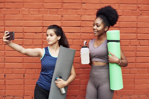 Image de jeunes femmes athlétiques étonnantes prenant un selfie contre un mur de briques.