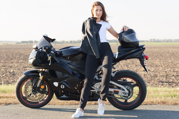 Image de jolie femme porte un t-shirt blanc, un pantalon noir et des baskets blanches, s'assoit sur une moto, garde la main sur le casque