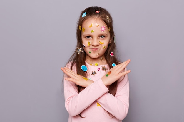 Image d'une jolie petite fille aux cheveux bruns recouverte d'autocollants posant isolé sur fond gris a croisé ses mains en disant qu'il n'y a aucun moyen de montrer un signe d'interdiction