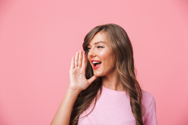 Image de joyeuse femme européenne avec de longs cheveux bruns bouclés regardant de côté à copyspace et en criant ou en appelant quelqu'un, isolé sur fond rose