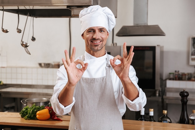 Image de joyeux chef masculin en uniforme blanc souriant, debout à la cuisine au restaurant