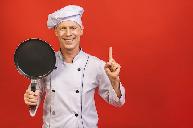 Image de joyeux chef principal homme en uniforme de cuisinier souriant et tenant la poêle à frire isolé sur fond de mur rouge.